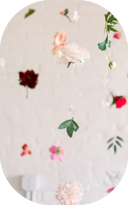 A soft, dreamy scene featuring various flowers suspended in the air, gently hanging against a white brick background. The flowers include soft pink peonies, bright red blooms, and green leaves, creating a floating floral display.
