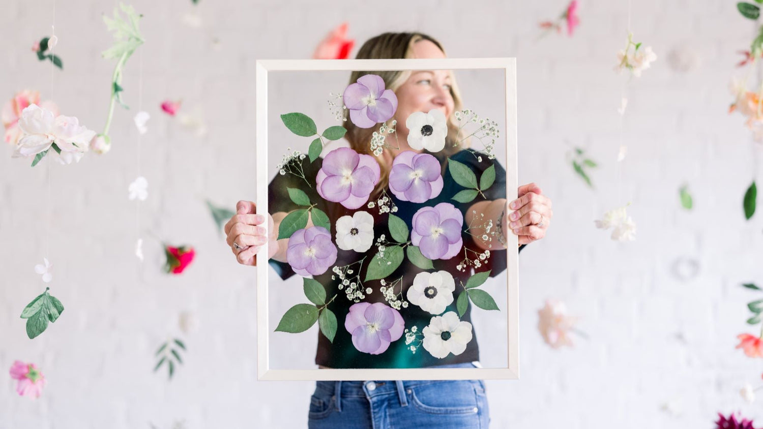 Amy, the founder of A Home in Bloom, holding a framed piece of pressed flower art featuring purple and white flowers with green leaves. Behind her, colorful flowers are suspended in the air against a soft white brick background.