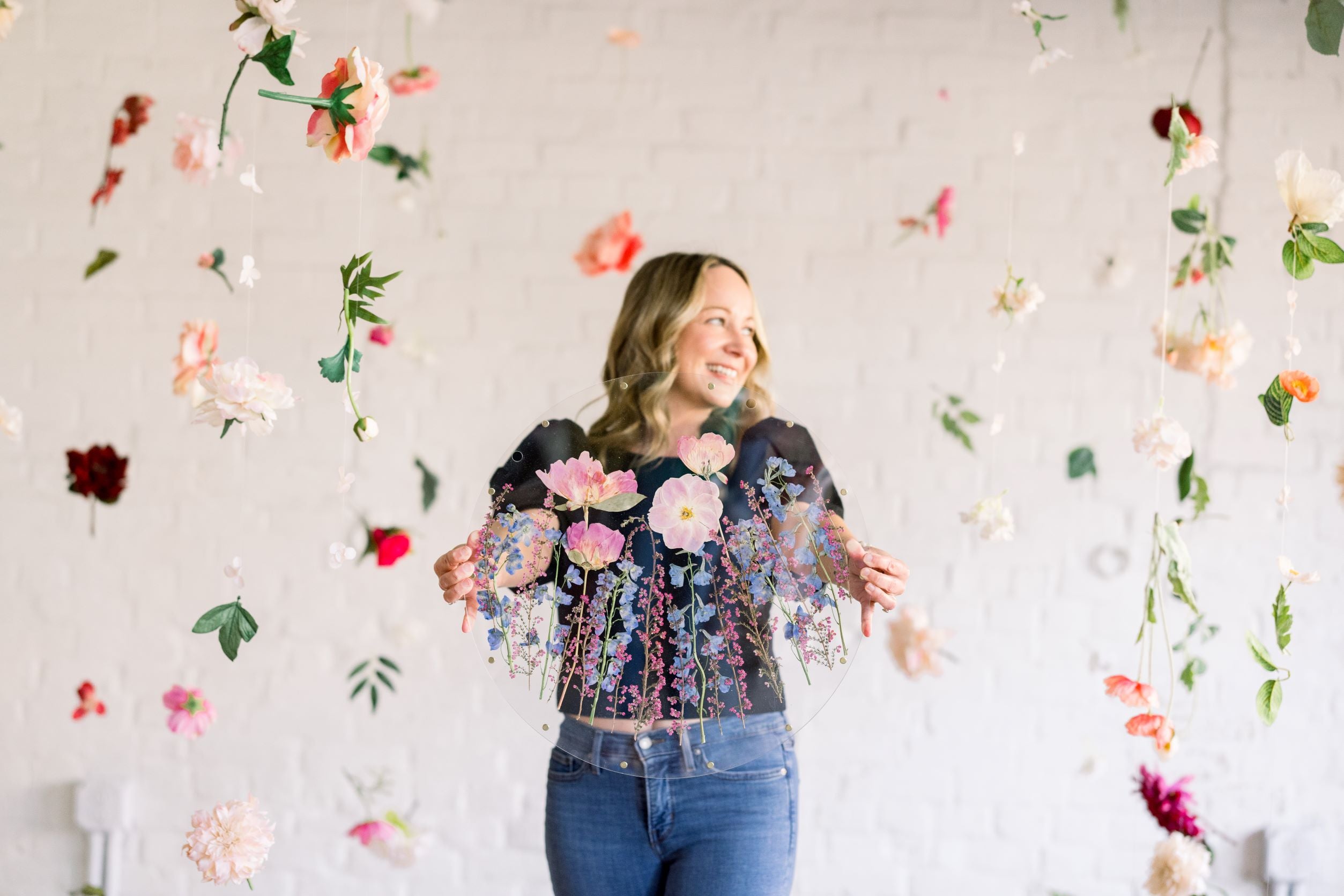 Amy, the founder of A Home in Bloom, standing in front of a white brick wall with colorful flowers hanging around her. She is holding a circular resin piece featuring pressed pink and purple flowers, including poppies and violets, with delicate greenery. She is smiling and appears joyful, surrounded by a beautiful, floral backdrop.