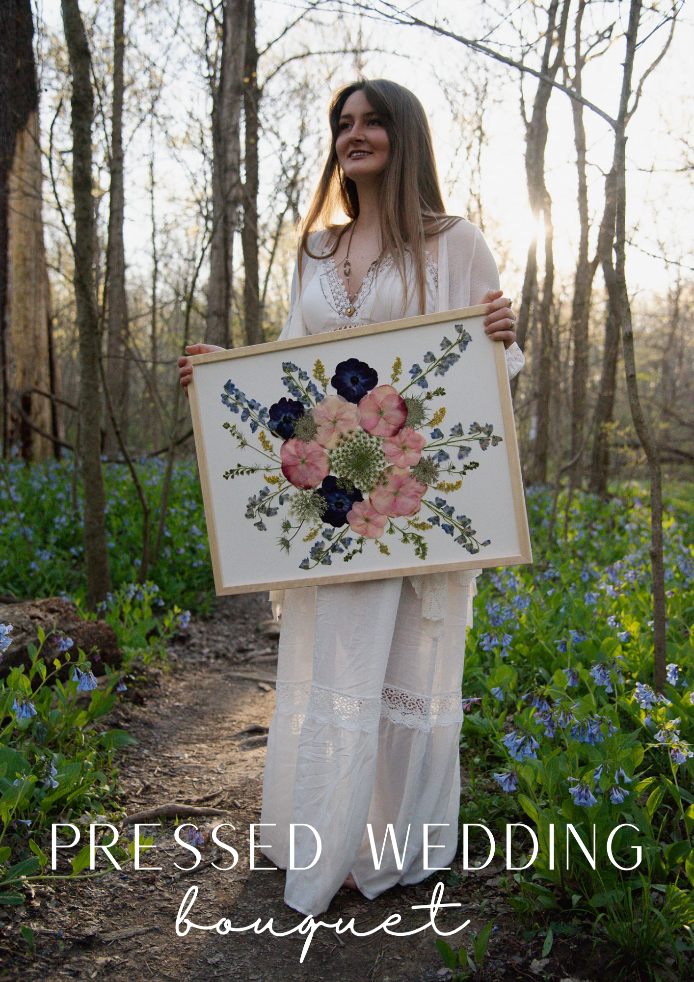 This image features a woman wearing a white boho dress, standing outdoors in a wooded area while holding a framed pressed flower bouquet. The bouquet, displayed in the frame, is a colorful arrangement featuring soft pink and deep blue flowers surrounded by greenery. The text at the bottom of the image reads "Pressed Wedding Bouquet," suggesting the preservation of a wedding bouquet through pressing flowers into a custom-designed piece.