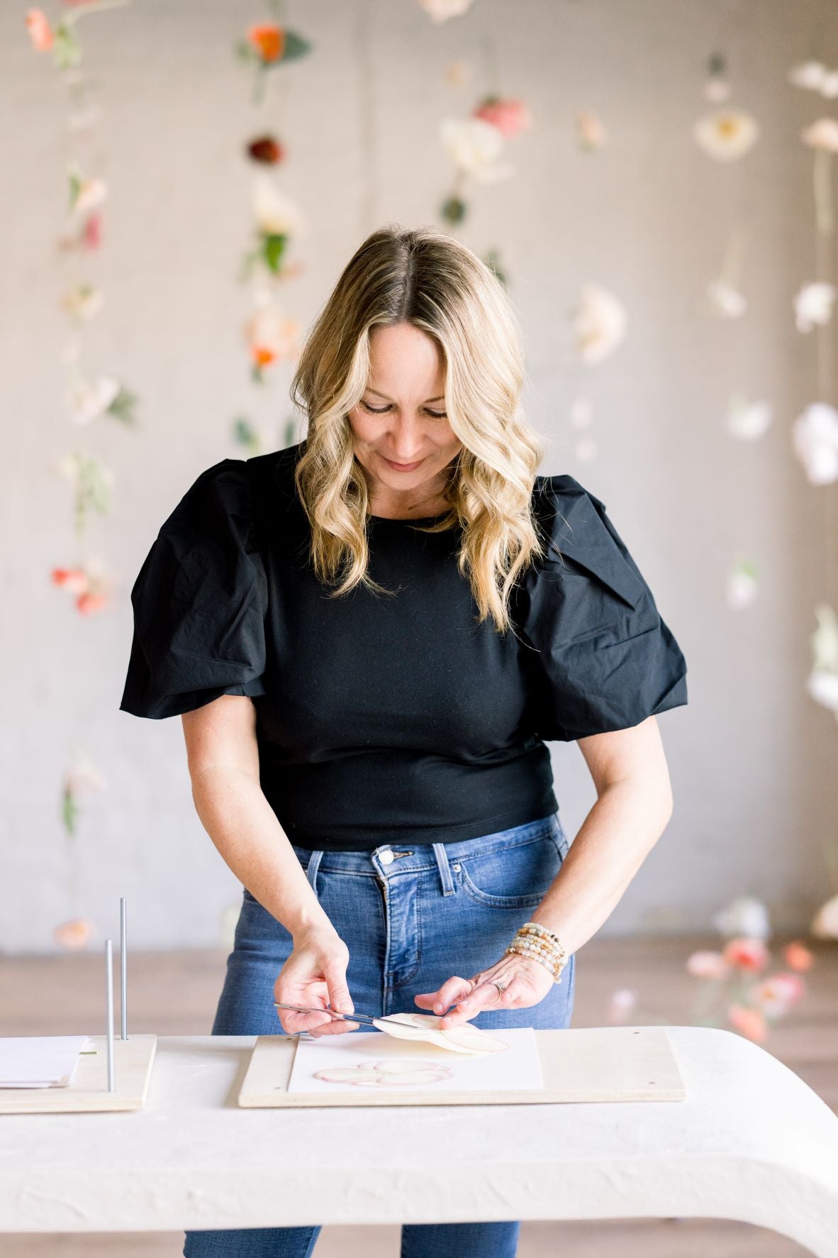 Amy, the founder of A Home in Bloom, standing at a table in the studio, focused on arranging pressed flowers. She is wearing a black top and jeans, with delicate floral arrangements suspended in the background. Amy is holding a flower petal and carefully placing it on a canvas as she works.