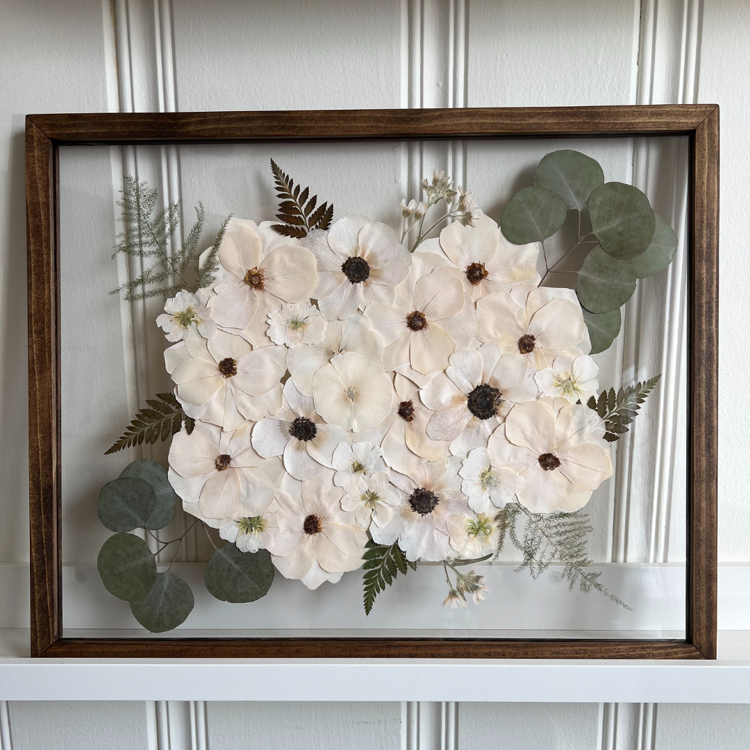 A framed piece of pressed flower art featuring a soft arrangement of white anemones with dark centers, complemented by eucalyptus leaves, ferns, and small white flowers. The artwork is beautifully displayed in a wooden frame, with the flowers arranged in a symmetrical pattern.