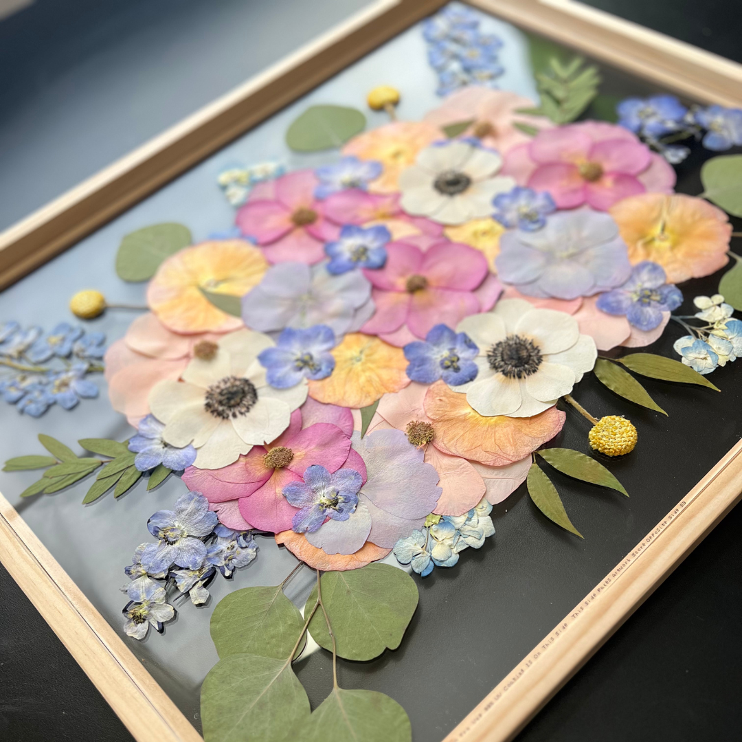 A framed piece of pressed flower art featuring a vibrant mix of pink, purple, and yellow flowers, including anemones, pansies, and violets, along with delicate greenery. The arrangement is carefully placed on a dark background, and the frame is light wood.