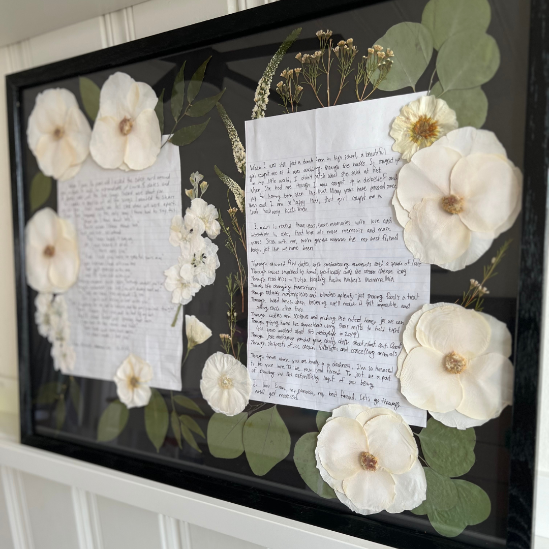 A framed piece of artwork featuring wedding vows written on two sheets of paper, surrounded by delicate white pressed flowers, including large blooms and greenery. The soft floral arrangement adds a personal touch to the handwritten vows, which are elegantly framed against a dark background.