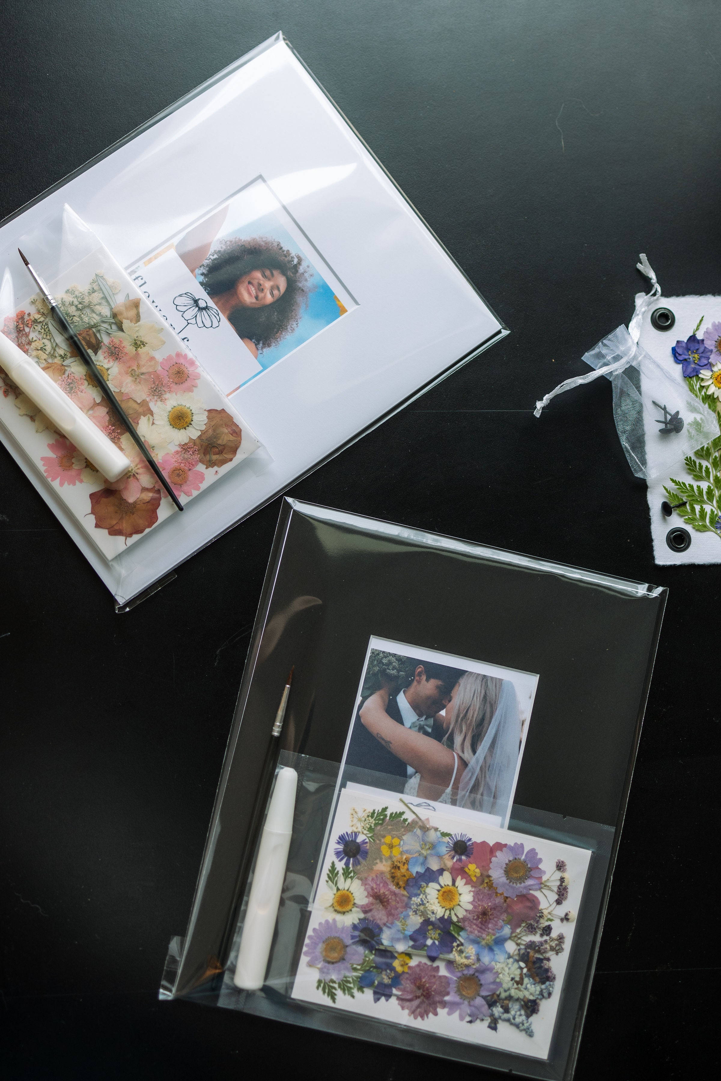 Two DIY pressed floral kits, each with a photo frame and flowers ready to be arranged. The top kit includes a photo of a person with a floral envelope containing pressed flowers, a brush, and a glue stick. The bottom kit features a photo of a couple and a collection of colorful pressed flowers, with a brush and adhesive pen for assembling the design.