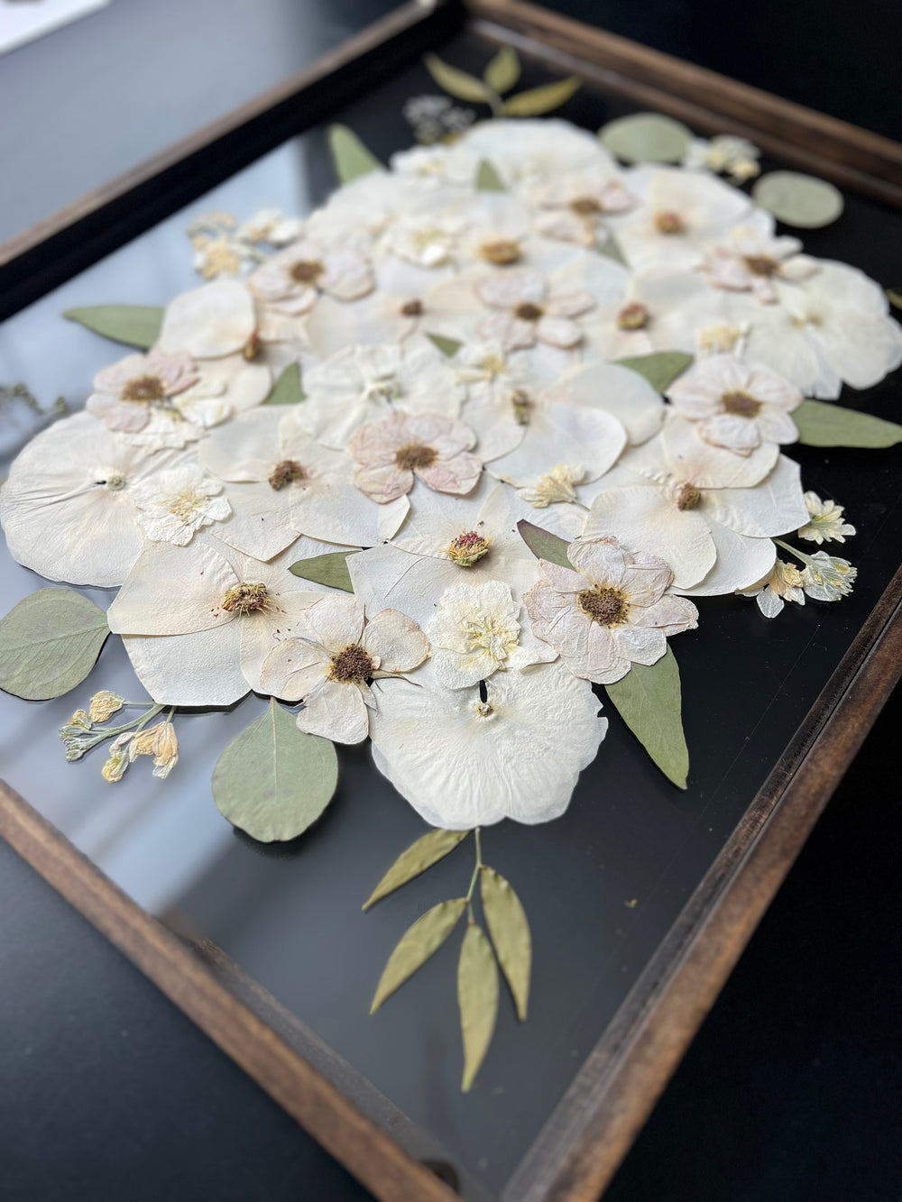 A close-up of a beautifully arranged pressed flower art featuring delicate white and pale pink flowers, leaves, and soft greenery.