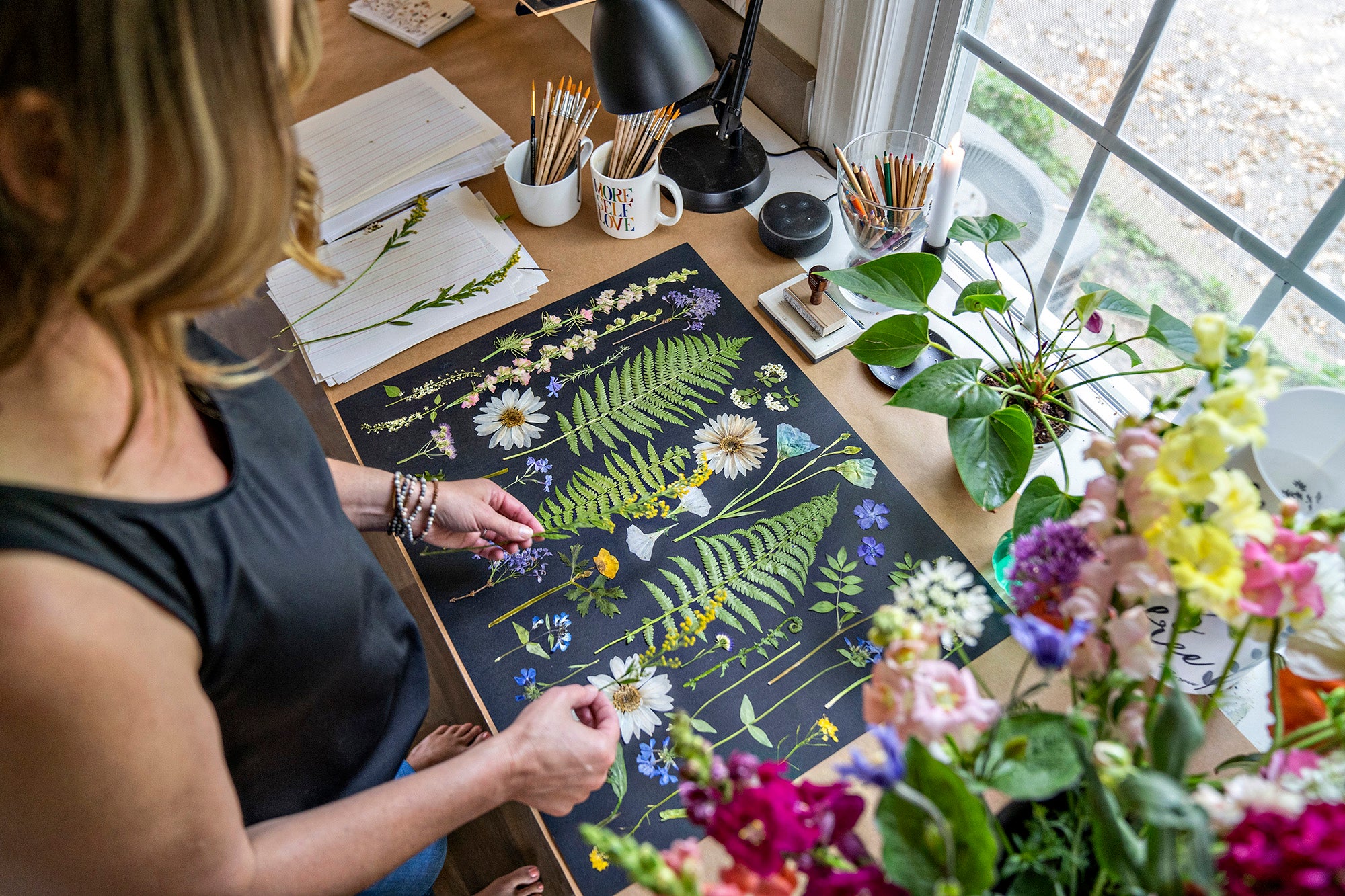 Amy arranging pressed flowers on a black background in a cozy, bright workspace. The table is filled with colorful flowers, art supplies like brushes and pens, and a potted plant. The person is carefully placing flowers, showcasing a creative floral art project.